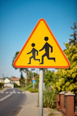 Karşıya geçen çocuklar için üçgen şeklinde bir uyarı işareti. Açık mavi gökyüzüne karşı çekilen fotoğraflar, sürücülere okul ve yaya alanlarının yakınında yavaşlamalarını hatırlatıyor..