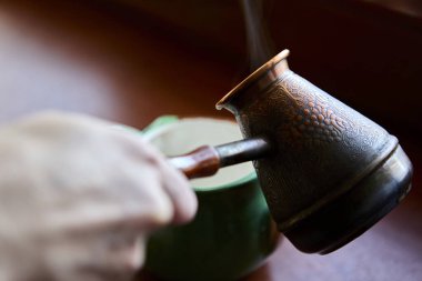 Kahve bakır bir kafesten, ahşap bir pencere pervazına yerleştirilmiş yeşil bir seramik kupaya dökülüyor. Gün ışığında buhar yükselir, sıcak sıcaklık katar..