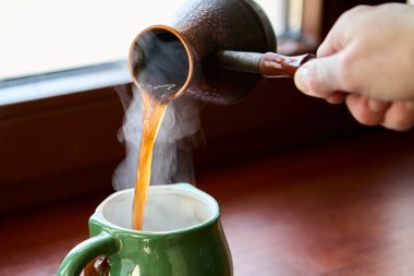 Sıcak koyu kahve, ahşap pencere pervazına seramik bardağa dökülüyor. Doğal ışık rahatlatıcı bir içecek havası yaratır..