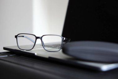 Yumuşak doğal ışıkla aydınlatılan bir masa yüzeyindeki kalem ve gözlük kutusunun yanındaki kapalı bir dizüstü bilgisayarın üzerine yerleştirilmiş şık siyah gözlükler. Çalışma alanı elementleriyle modern natürmort.