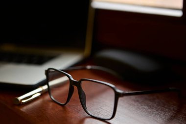 Altın bir kalem ve dizüstü bilgisayarın yanındaki parlak ahşap bir masada dinlenen okuma gözlüklerinin yakın çekimi. Kompozisyon, şık bir profesyonel ofis ya da ev çalışma alanı ortamı yaratır.
