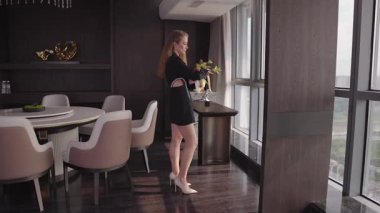 Siyah ceketli şık genç bir kadın kollarını kavuşturmuş modern lüks bir şekilde pencereden dışarı dikkatlice bakıyor. Zarif ve minimalist bir dekor ve yumuşak gün ışığı.