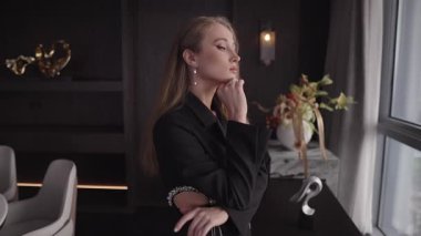 Siyah ceketli düşünceli bir kadının, lüks bir iç mekanda pencerenin yanında dikilirken yakın çekimi. Eli, zarif modern dekorlar ve hafif ışıklandırmalarla çevrelenmiş, dalgın dalgın aşağı bakarken çenesinin altında duruyor.