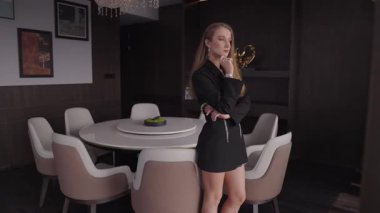 Siyah ceketli bir kadın modern bir dairede yuvarlak bir yemek masasının yanında duruyor. Elini çenesine koyar, saatine göre kontrol eder. Kamera, kusursuz bir geçiş yaratmak için duvara bir itme ile biter. Ruh hali hüzünlü ve dalgındır..