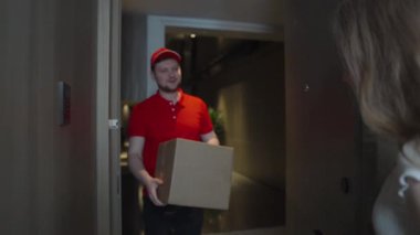 Kırmızı üniformalı bir kurye kapıdaki müşteriye karton bir kutu verir. Kapalı teslim sahnesinde e-ticaret, lojistik hizmetleri ve modern nakliye kolaylığı vurgulanıyor.