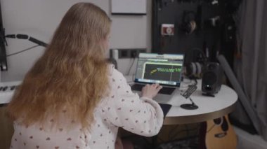 Dizüstü bilgisayarda çalışan kadın müzik yapımcısı, kıvırcık saçlı, profesyonel ses düzeneği ve çalışma alanını çevreleyen müzik aletleriyle ev stüdyosunda izler yaratıyor.