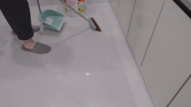 Siyah pantolon ve terlik giyen profesyonel temizlikçi faraş ve süpürge kullanarak beyaz fayanslı zemindeki toz ve kiri düzenli olarak süpürür.