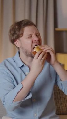 Kanepede oturan bir adamın yavaş çekim sahnesi, bir hamburgerin her ısırığını zevkle ve kapalı gözlerle yiyor, rahat bir ev ortamında.