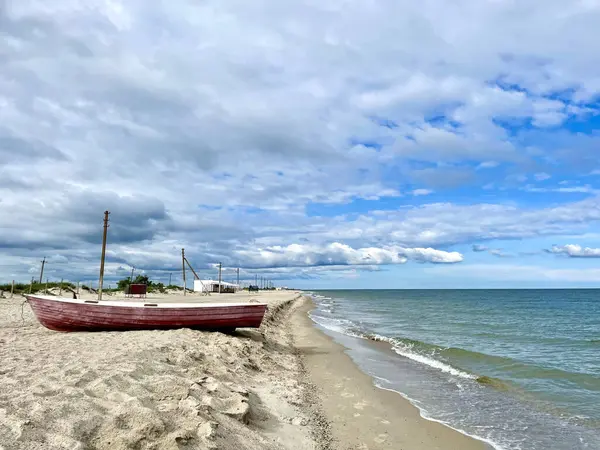 Kırsal, kırmızı ahşap bir kayık kumlu bir sahilde parçalı bulutlu bir gökyüzünün altında denizin sakin, turkuaz sularının yanında duruyor. Arka planda, elektrik direkleri kıyı şeridi boyunca uzanıyor, bu da uzak ya da seyrek gelişmiş bir bölge olduğunu gösteriyor..