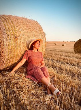 Dişi çiftçi ya da tarım işçisi hasat edilen buğday tarlasında saman balyasına yaslanarak mola veriyor. Altın saat ışığı samanı ve kırsal araziyi aydınlatıyor.