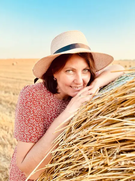 Çiçekli elbise ve güneş şapkalı bir kadının geniş saman balyalarıyla bir tarlada rahatlarken tam boy fotoğrafı. Açık mavi gökyüzü ve sıcak akşam ışığı Ukrayna kırsalında yaz mevsimi manzarası yaratıyor.