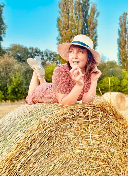 Dişi çiftçi ya da tarım işçisi hasat edilen buğday tarlasında saman balyasına yaslanarak mola veriyor. Altın saat ışığı samanı ve kırsal araziyi aydınlatıyor.