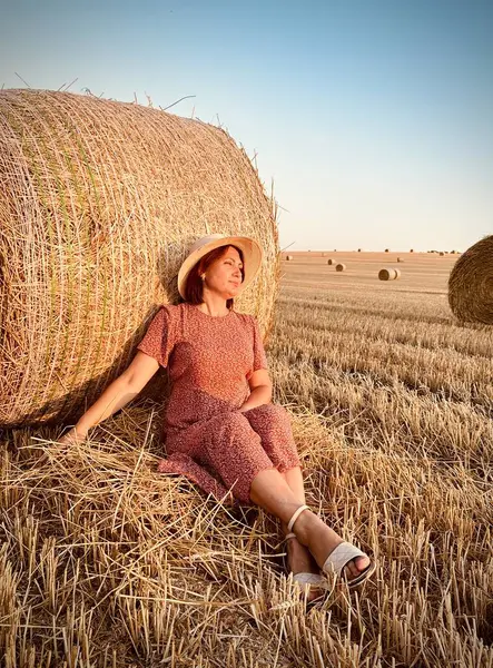 Dişi çiftçi ya da tarım işçisi hasat edilen buğday tarlasında saman balyasına yaslanarak mola veriyor. Altın saat ışığı samanı ve kırsal araziyi aydınlatıyor.
