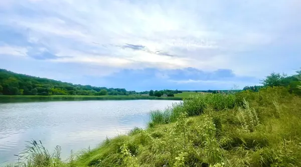 Berrak yaz manzarasında yeşil ağaçlar ve ön planda uzun otlarla çevrili sakin bir göl vardır. Karşı kıyıda, sakin suya yansıyan küçük bir ahşap kulübe görülebilir. Gökyüzü yumuşak bulutlar ve gölgelerle süslenmiş.