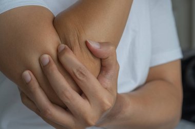 Asyalı bir adam dirseğini tutar, eklem ya da tendon rahatsızlığını gösterir. Beyaz gömleği ve yumuşak arkaplanı klinik ama ilişkilendirilebilir bir ortam yaratıyor. Sağlık ve Tıp Konsepti.