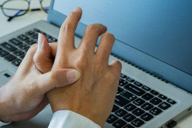 Asyalı bir adam, ofis sendromunun erken belirtileri üzerinde çalışırken başparmak rahatsızlığı gösterir. Yanındaki bardaklar uzun ekran görüntülerine işaret ediyor. Sağlık ve Tıp Konsepti.