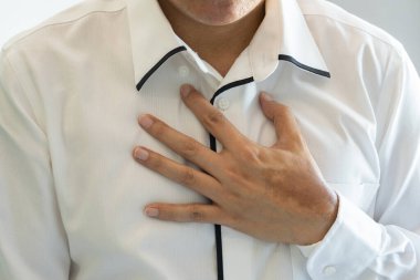 Asyalı bir adam göğsüne endişe ile dokunuyor, muhtemelen rahatsızlık veya kalp rahatsızlığı belirtileri gösteriyor. Tarafsız ve yumuşak bir ortamda beyaz bir gömlek giyiyor. Sağlık ve Tıp Konsepti.