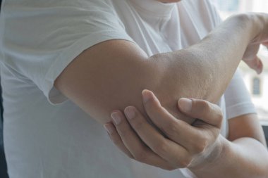 Asyalı bir adam sağ dirseğini görünür bir gerilimle tutar ve lokal ağrı ya da gerginliğe işaret eder. Yumuşak ışıklandırılmış arka plan bu rahatsızlığın tıbbi ifadesini çerçeveler. Sağlık hizmetleri tıbbi konsepti.