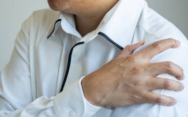 Asyalı bir adam sol omzunu tutar. Muhtemel kas veya eklem ağrısına işaret eder. Tarafsız bir arka plan, kapalı ve klinik olmayan bir ortama işaret ediyor. Ofis sendromu kavramı.