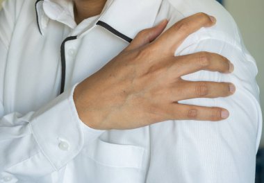 Asyalı bir adam sol omzunu tutar ve lokal ağrı ya da kas dokusunun gerildiğini belirtir; günlük aktivitelerle ilgili bir durum olduğunu belirtir. Sağlık ve Tıp Konsepti.