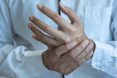 Asyalı bir erkek, olası eklem ağrısı veya kas iskeleti rahatsızlığını belirtmek için sol elini tutar; ortam nötr ışıklandırmayla klinik veya ev bazlı görünür. Sağlık ve Tıp Konsepti.