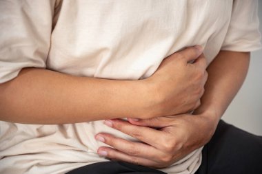 Karın bölgesini kavrayan kişi, mide ağrısı, şişkinlik ya da iltihaplanmadan kaynaklanan karın ağrısına işaret ediyor. Kapalı alan ayarları bir ev ya da klinik bağlam içerir.