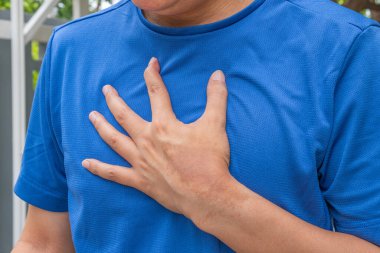 Asyalı bir adam antremandan sonra endişeyle göğsünü tutuyor. Arka planda rahatsızlık verici spor salonu belirtileri gösteriyor. Kalp hastalıkları, solunum sorunları veya kalp riski üzerine sağlık reklamları için ideal.