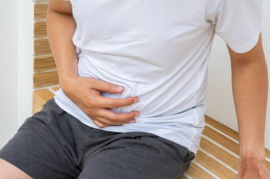 Asyalı adam verandada oturmuş idman sonrası kramplardan karnını sıkıyor. Sindirim rahatsızlığı ve düzgün bir sağlık yönetimi ihtiyacını gösteriyor.