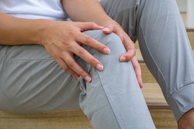 Asyalı adam ahşap basamaklara oturmuş, dizini sıkıca tutmuş, koşma sonrası rahatsızlığı, muhtemel dizkapağı gerginliği ya da eklem iltihabı. Sabah egzersizi, spor sakatlığı, rehabilitasyon, sağlık..