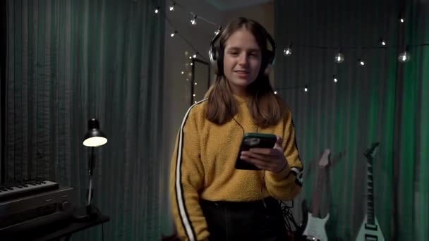 ヘッドフォンの美しい女の子は 掃除機の部屋を掃除し スマートフォンで表示します 黄色のセーターと手フーバーの女性は部屋をきれいに — ストック動画