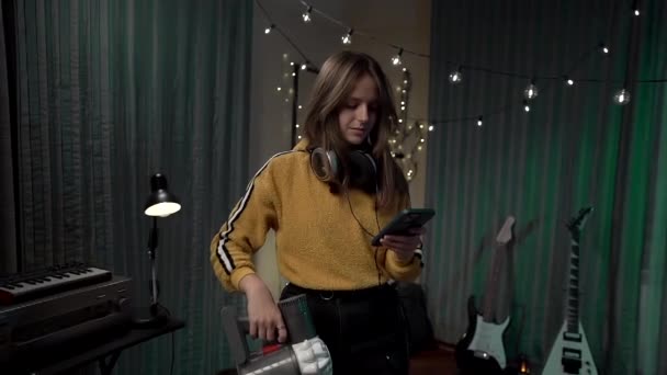 ヘッドフォンの美しい女の子は 掃除機の部屋を掃除し スマートフォンで表示します 黄色のセーターと手フーバーの女性は部屋をきれいに — ストック動画