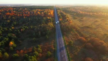 Hava manzaralı insansız hava aracı, güneşli bir sonbahar gününde, kırsal köy yolunun üzerinde, sinematik, canlı, renkli ormanlarla çevrili bir şekilde gidiyor..