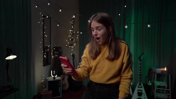 自宅で黄色のセーターでデバイス上の感情ガールハッピーチェックアプリ レッドスマートフォンを見て勝つ入札を祝う若いユーフォリック女性 — ストック動画