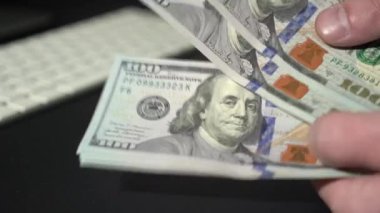 Erkek eli para sayıyor. Adam arka plandaki monitörlerde ticari grafiklerle 100 Amerikan doları hesaplıyor. Finans ve zenginlik kavramı