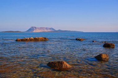 İtalya 'nın Sardunya kıyılarından görülen Tavolara adasının manzaralı deniz manzarası. Ön planda görünen kayalar ve berrak mavi gökyüzünün altındaki uzak dağ manzaralı sakin Akdeniz suları. Seyahat, turizm, doğa için ideal.