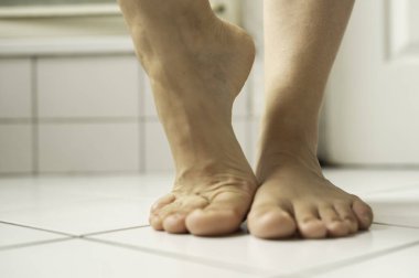 Çıplak ayak, parlak beyaz banyo fayanslarına basarak doğal gün ışığını yansıtıyor. Hareketi ve kapalı ortamın temizliğini vurgular.