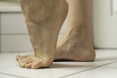 Çıplak ayak, beyaz banyo fayanslarında parmak ucunda, gün ışığıyla aydınlatılmış. Temiz bir iç mekanda ince bir hareket ve deri ayrıntısı yakalar.