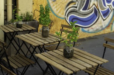 Parisli canlı bir kafe terası, sıcak bir yaz gününde misafirler için hazır. Kavanozlarda saksı bitkisi olan ahşap masalar renkli bir grafiti duvarına karşı duruyor, dışarıda yemek yiyor..