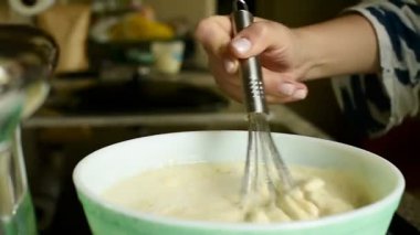 Bir kadının elini paslanmaz çelik çırpıcıyla açık yeşil bir kasede karıştırdığını gösteren HD video çerçevesi. Konforlu bir ev mutfağında yemek hazırlama sürecini yakalar..