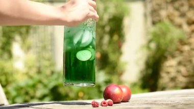 Bir kadının elleri sıcak bir yaz terasında canlı yeşil bir limonatayı karıştırır. Sahne taze nektarin ve ahududu bileşimiyle güçlendirilmiş, kırsal bir rahatlama hissi uyandırıyor. Tam HD.