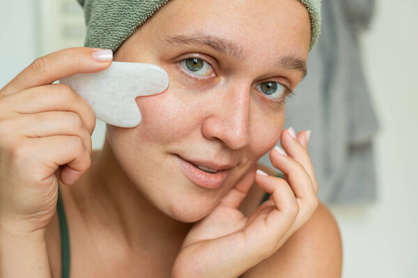 Молодая женщина с спокойным выражением лица использует белый гуа ша инструмент на ее лице. Мягкий естественный свет подчеркивает ритуал кожи, идеально подходит для красоты и оздоровительных тем.
