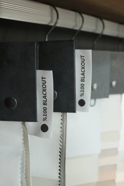 Sergi odasının korkuluklarında asılı% 100 karartma kumaş kumaş kumaş kumaş kumaş örneklerinin dikey yakın çekimi. Eyelevel görünümü, perde deposunda tam ışık bloğu kalitesini gösteren örnek klasörleri ve etiketleri gösterir