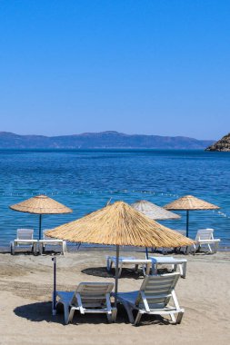 Beyaz plastik güneş şezlongları ve saman şemsiyeleriyle huzurlu kumlu bir sahil. Sakin turkuaz deniz ve açık mavi gökyüzünün altındaki uzak dağlar gevşemeyi ve yaz tatili titreşimlerini çağrıştırıyor. Erdek. Türkiye