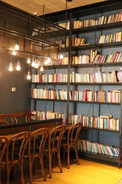Renkli kitaplarla dolu uzun siyah metal bir kitaplık, asılı ampullerle sıcak ışıklandırma ve uzun bir ortak masa etrafında dizilmiş ahşap sandalyeler içeren rahat bir kütüphane köşesi. Okumak ya da sessiz çalışmak için mükemmel bir yer.