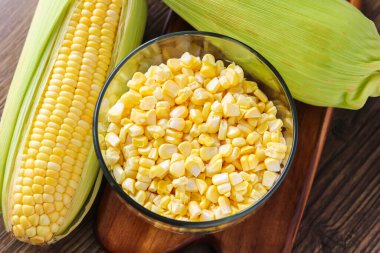 A view of two fresh ears of corn, one partially husked, next to a clear glass bowl of sweet corn kernels, all arranged on a wooden cutting board.