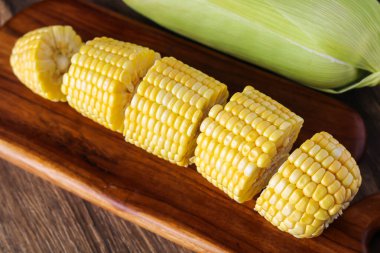 An unhusked ear of corn with several cut pieces of corn on the cob, arranged on a wooden cutting board.
