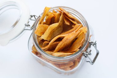Closeup of a glass jar filled with crispy banana chips. Captured with natural light on a white background.