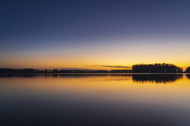 Hollanda 'nın Alkmaar kenti yakınlarındaki sakin bir gölün üzerinde sakin bir günbatımı. Gökyüzü turuncu ve pembe yumuşak renklere bürünüyor. Sakin sulara ve kırsal kesime nazikçe yansıyor.