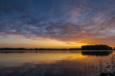 Hollanda 'nın Alkmaar kenti yakınlarındaki sakin bir gölün üzerinde sakin bir günbatımı. Gökyüzü turuncu ve pembe yumuşak renklere bürünüyor. Sakin sulara ve kırsal kesime nazikçe yansıyor.