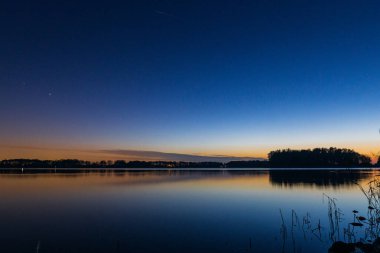 Hollanda 'nın Alkmaar kenti yakınlarındaki sakin bir gölün üzerinde sakin bir günbatımı. Gökyüzü turuncu ve pembe yumuşak renklere bürünüyor. Sakin sulara ve kırsal kesime nazikçe yansıyor.